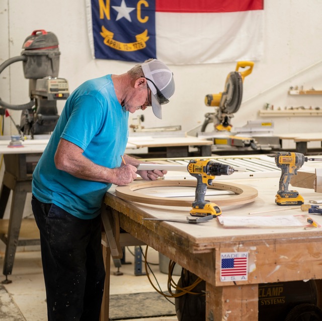 A craftsman hand-building a custom shutter at the Artisan / A Shade Above workshop in Monroe, NC, under the NC and US flags.